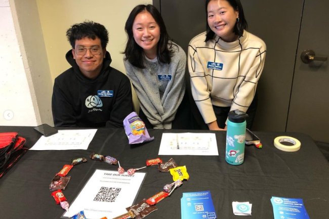Three of our staff smiling warmly and enthusiastically from behind a table at our first Volunteer Fair. They made the table decorative, with a heart of candy around one of our flyers.